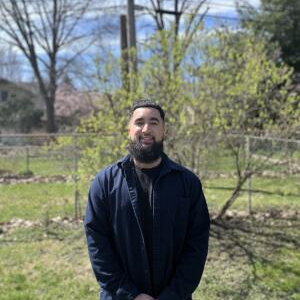 A man stands, hands clasped, outside in a nature space.